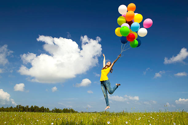 Happy young woman holding colorful balloons and flying over a green meadow
